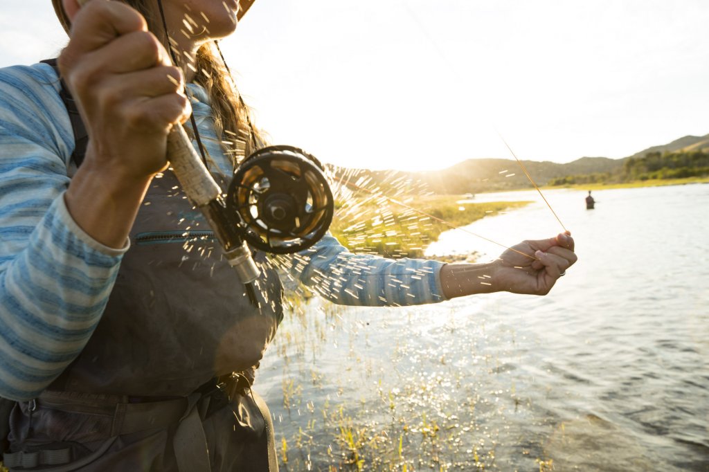 A close up of a person fly-fishing in a river, holding a fishing pole in one hand and tugging the line with the other.