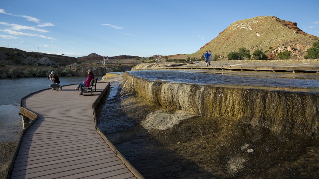 One traveler kneels to take a photo while another lounges in a chair, two travelers walking in the distance behind them, at Hot Springs State Park in Thermopolis, Wyoming.