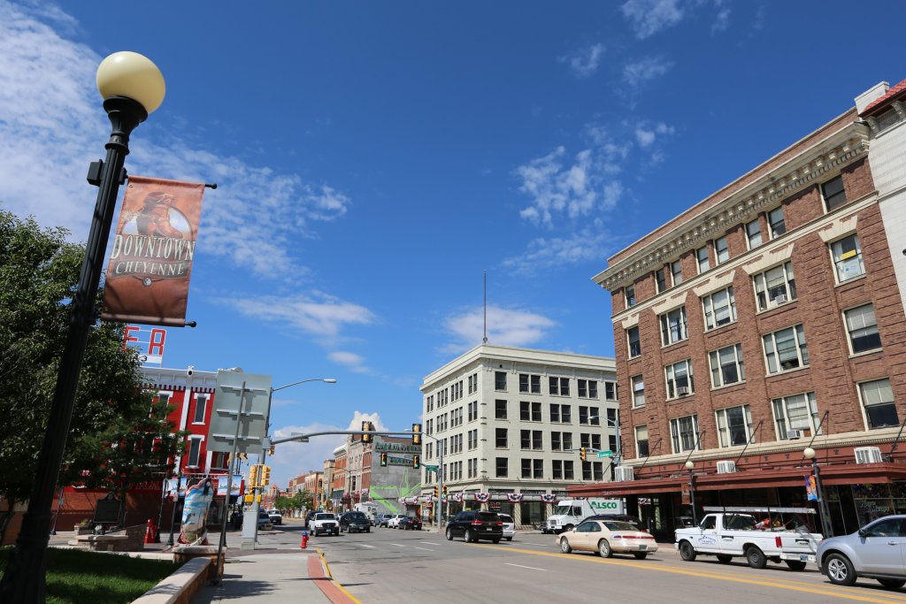 A colorful street view of bustling downtown Main Street in Cheyenne, Wyoming, where cars move along the street as a downtown sign hangs from above. 