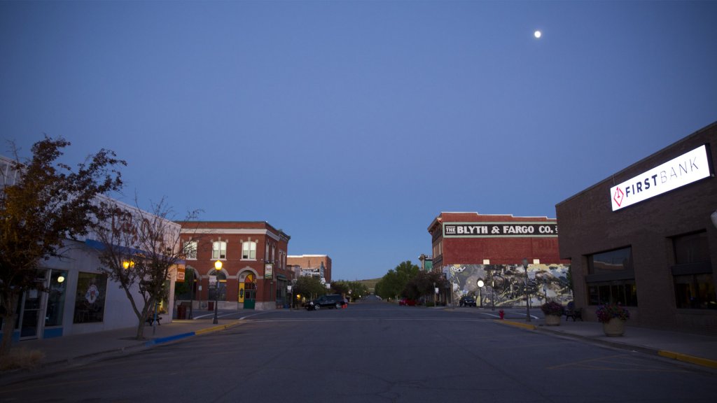 A night view of Main Street in Evanston, Wyoming, where the moon glows in an evening blue sky above a glowing First Bank sign on a building that's across from The Blyth & Fargo Co.