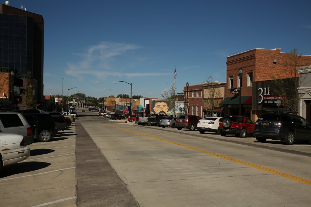 A view of the main street in Gillette, Wyoming, where the downtown area has cars that line the streets, including some in front of the 311 Restaurant & Lounge. In the distance is a painted mural of a cowboy with a mustache. 
