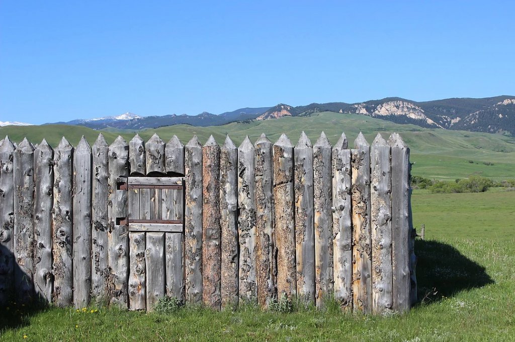 A sharpened log fence post sits before rolling green mountains near Fort Phil Kearny State Historic Site.
