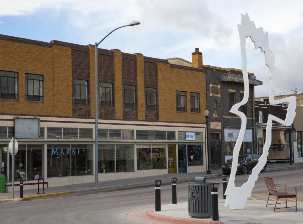 A view of the main street in Rollins, Wyoming, where a white statue of the outline of a bird soaring hangs before the building that reads, "Markey" with "Aflac" right next door. 