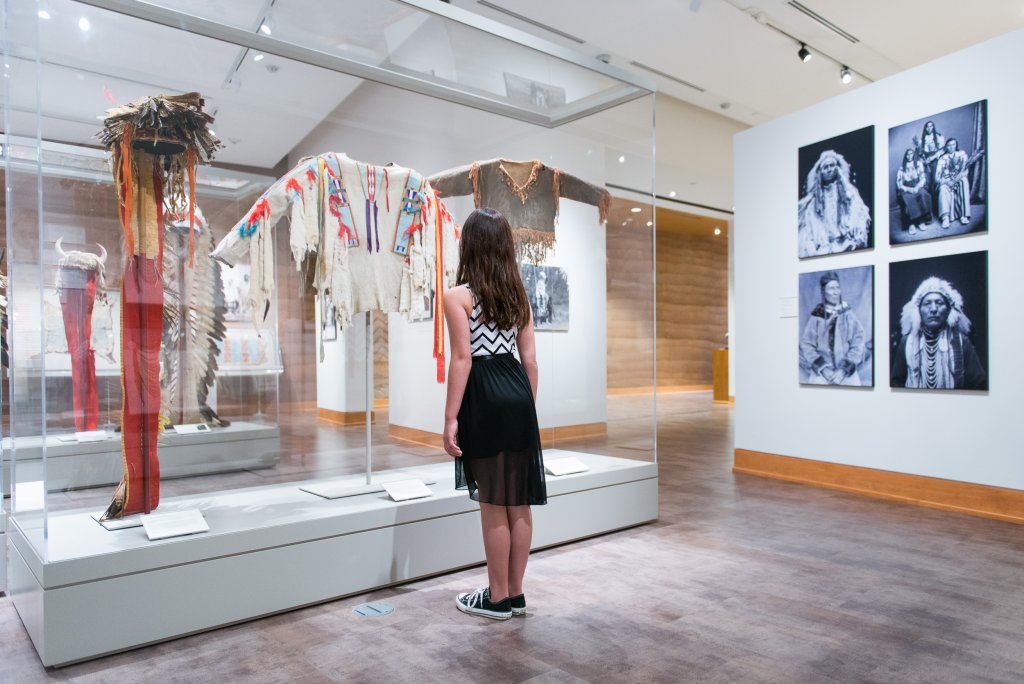 A woman looking at an exhibit of traditional Plains Indian clothing beside a wall of photographic displays at the Brinton Museum.