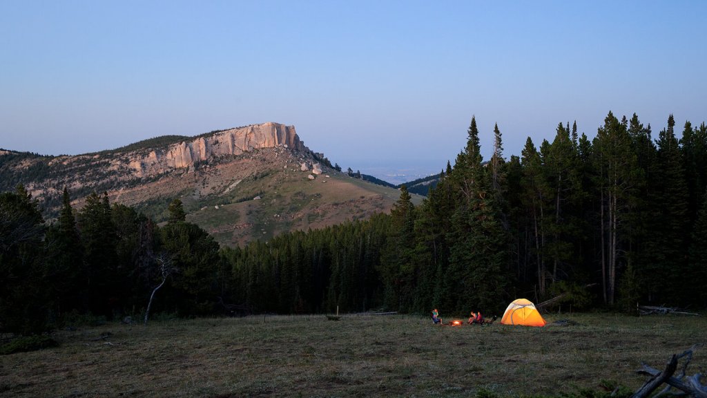 Campsite setup below mountainside as campers sit around an evening fire. 