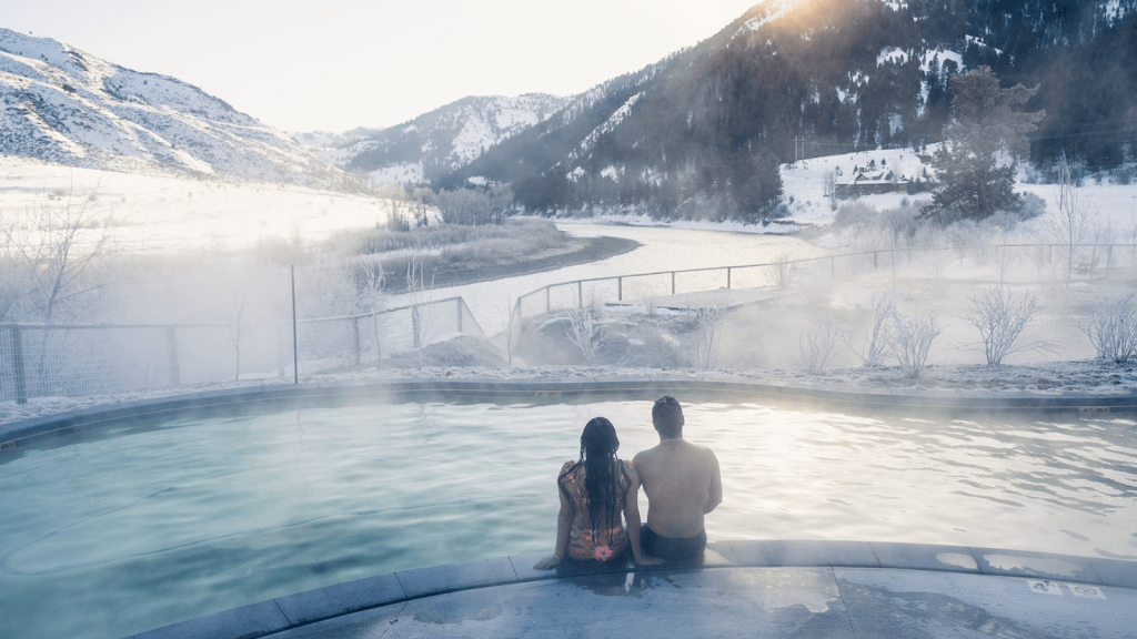 A couple sitting on the edge of a hot spring pool looking at the sun going behind the mountain.