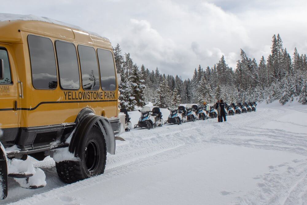 a yellow bus with snowmobiles behind it amongst a snowy white landscape in Yellowstone National Park during the winter. 