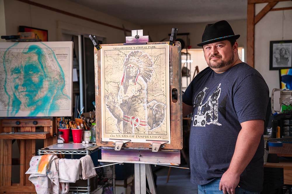 Robert Martinez stands next to his Yellowstone 150th commemorative art piece, which features Northern Arapaho leader Yellow Calf standing in the foreground and the image of an elk and a bison behind him. The image is overlaid on a map of Yellowstone National Park.