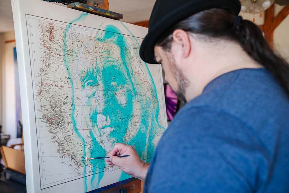 Robert Martinez stands at a canvas and paints an image of a Native American face over a map of the western United States.