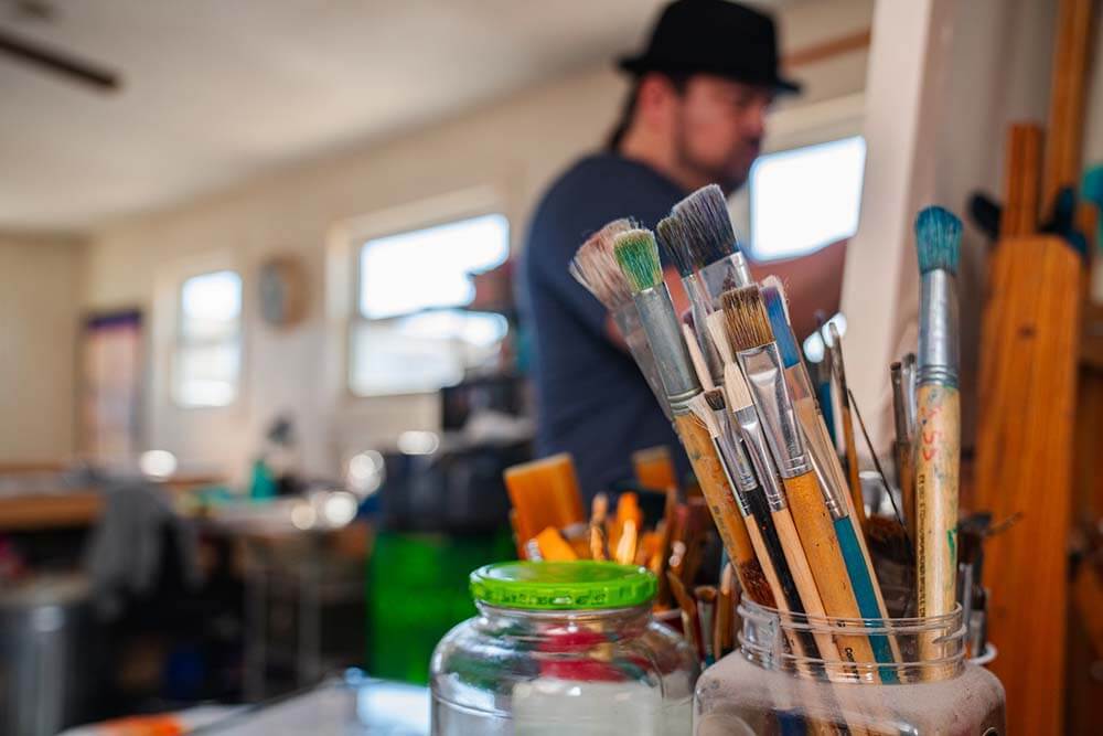 A set of different sized paint brushes sit in a jar at the focus, with artist Robert Martinez painting on a canvas in the background.