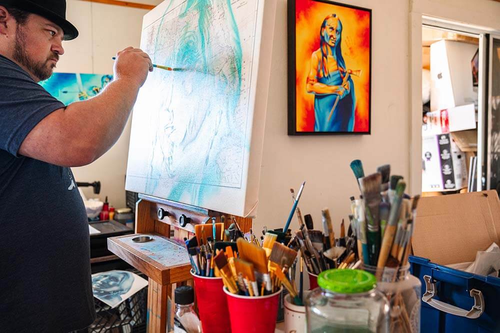 Artist Robert Martinez stands at an easel and paints the face of a Native American over a map. Cups and jars filled with paint brushes are on a table next to him. One of Martinez's completed paintings of a Native American hangs on a nearby wall.