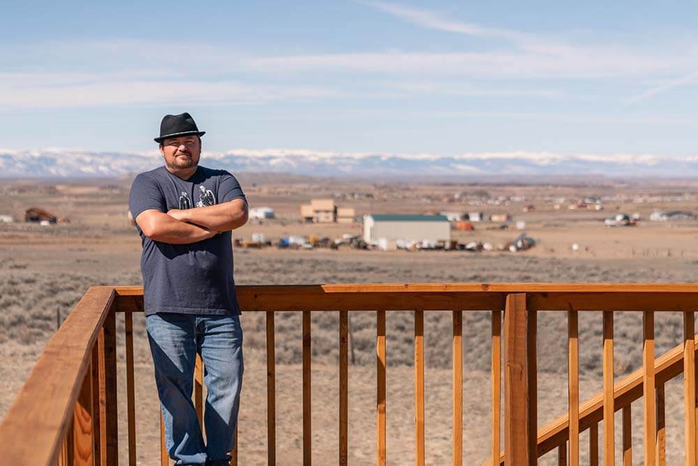 Artist Robert Martinez stands on a deck with his arms crossed and Wyoming's Wind River Range in the background.