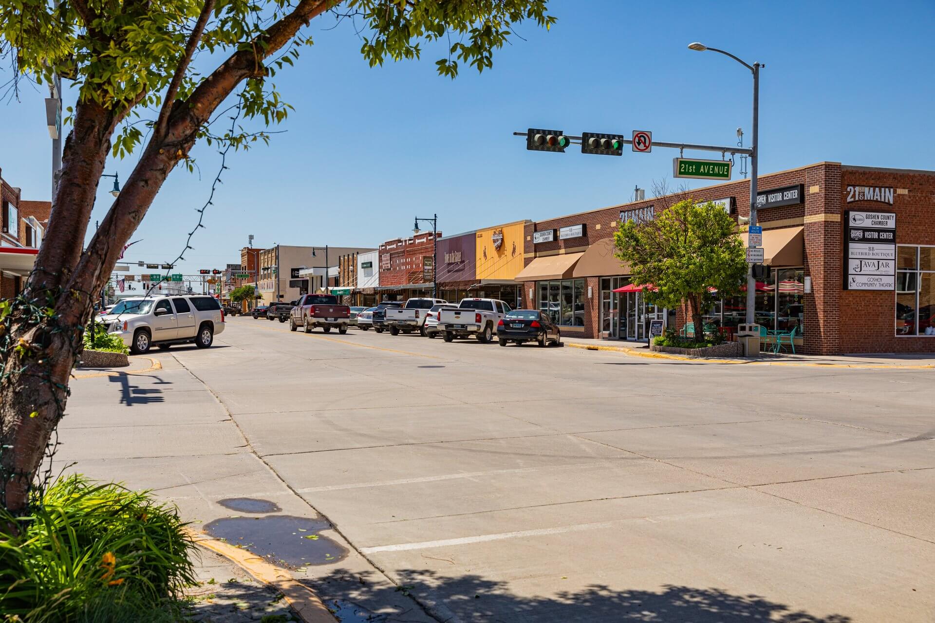 A view of the main street in Torrington, Wyoming where a building has a sign for 21st & Main next to the street sign for 21st Avenue as a tree leans to the right above the street on the left. 