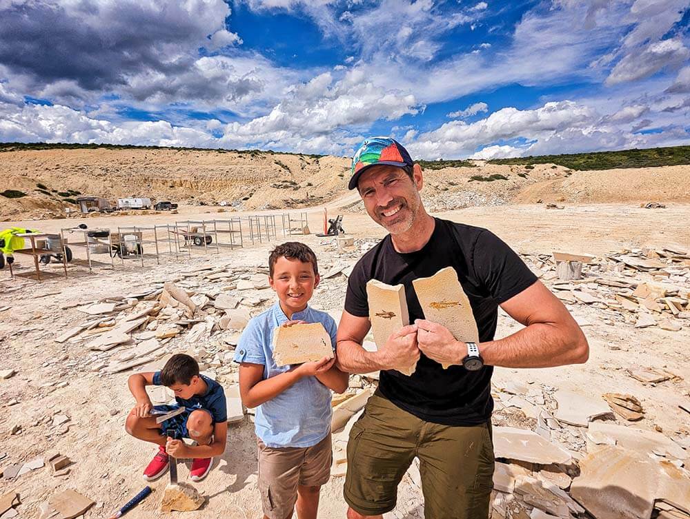 A father and son holding up fossils at the American Fossil Quarry as a boy digs for fossils in the background.