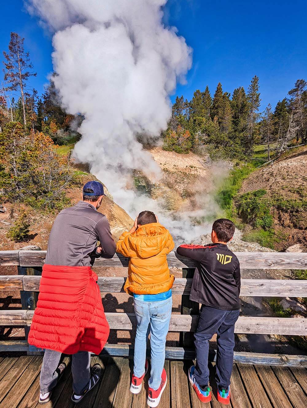 A father and two kids watch as steam erupts from a geyser.  