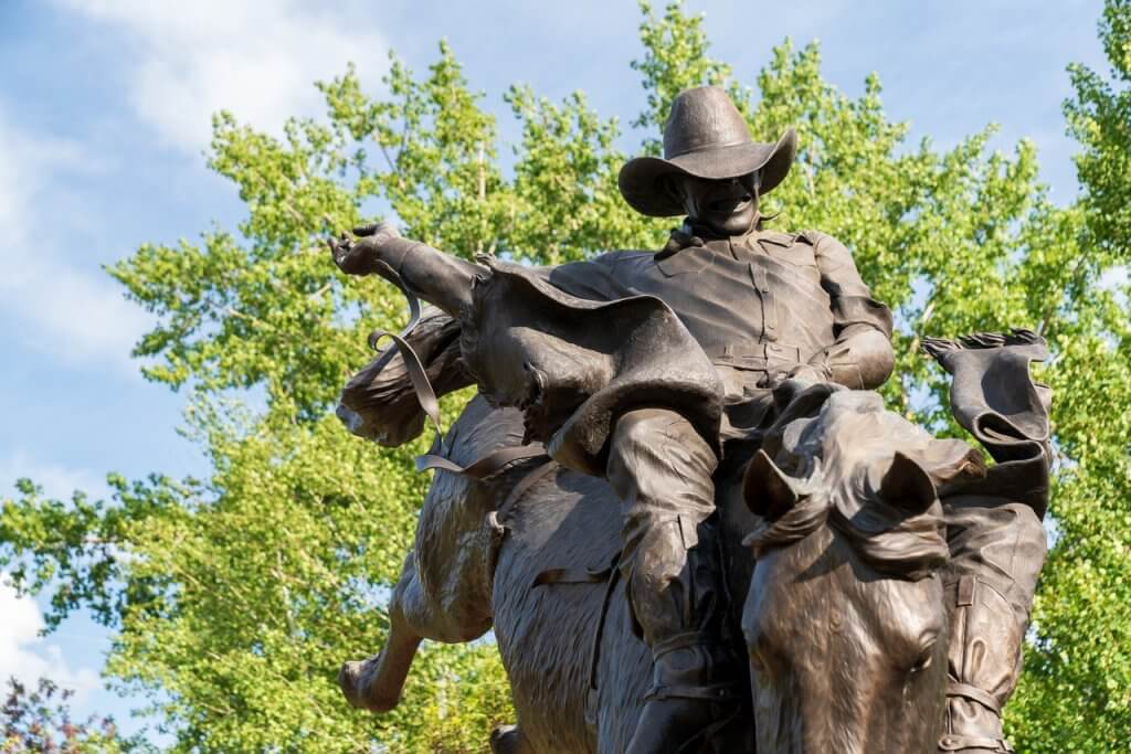 Bronze statue of Chris LeDoux, the renowned country singer and rodeo champion, dynamically portrayed in mid-ride on a bucking horse under a canopy of green leaves.