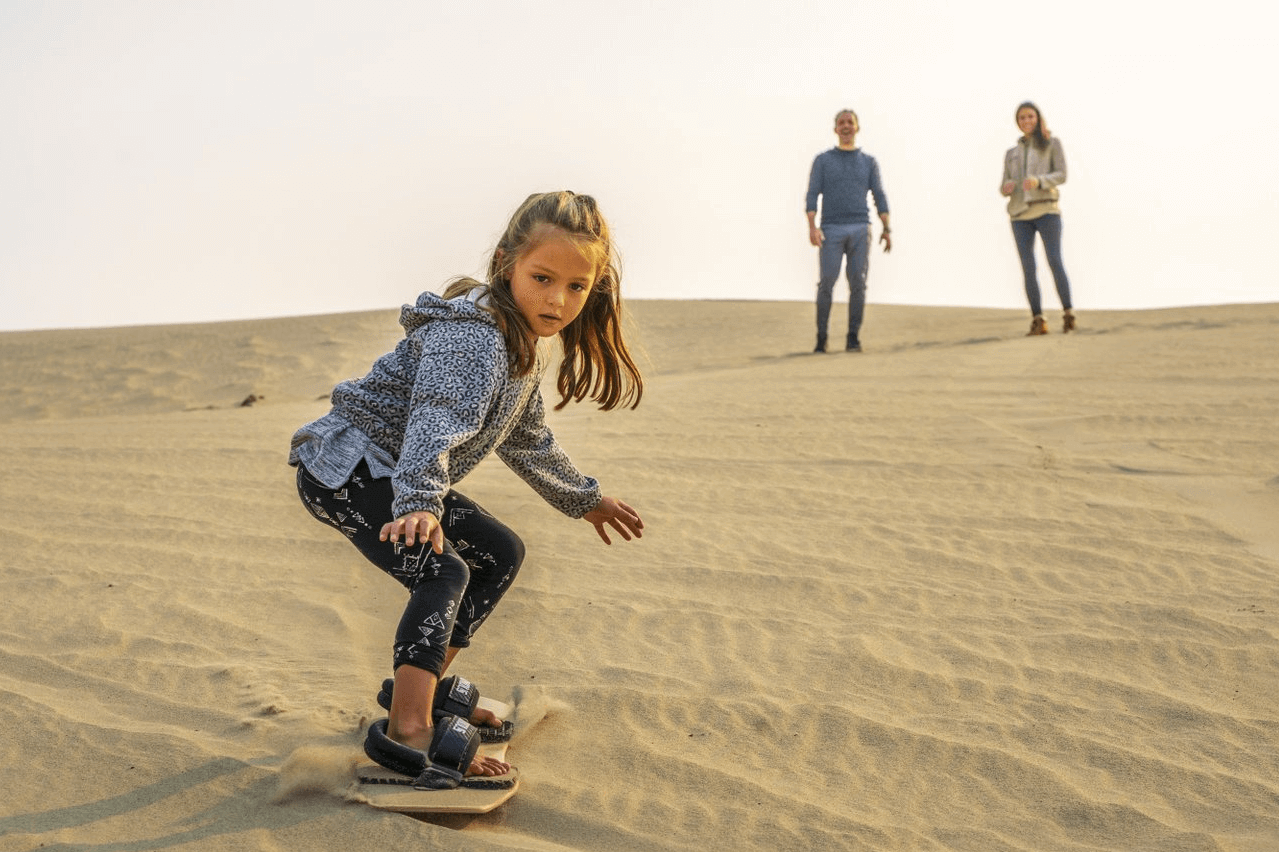 A young girl holds her hands out for balance as she sandboards across a sand dune as her parents watch from a distance.