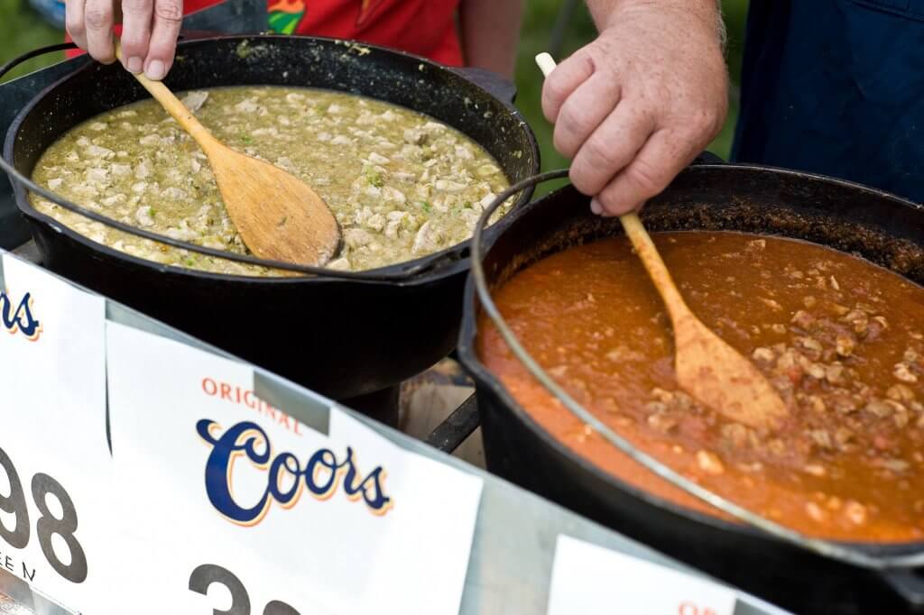 A close-up view of a person stirring a pot of vibrant green chili and pot of red chili at the Chugwater Chili Cook-Off.