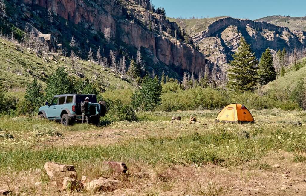 A person standing at the back of a Jeep a few yards away from a small tent within a valley surrounded tall trees and rock formations in the Shoshone National Forest.