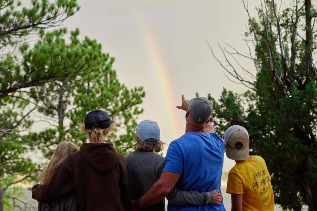 A family holds each other close as the father, who's wearing a baseball cap, points to a rainbow shining above them in the Wyoming sky.