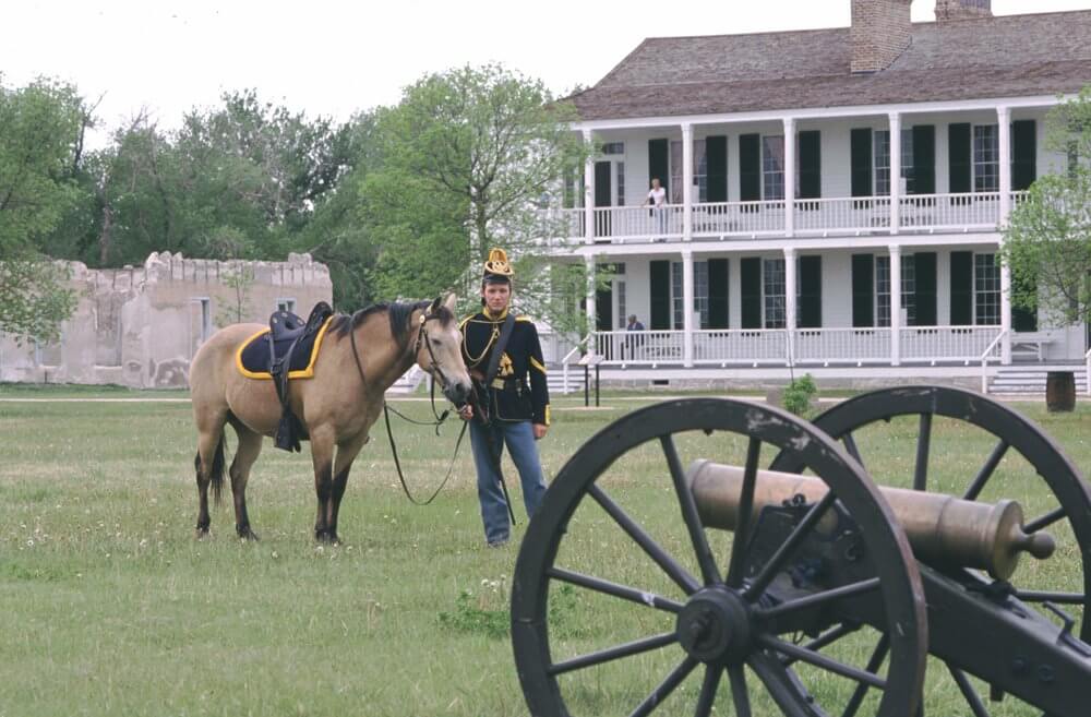Someone dressed as a soldier stands beside a horse in front of an old canon in the grass at the Fort Laramie National Historic Site. Behind them, a woman looks up from the second floor of a tall house. 