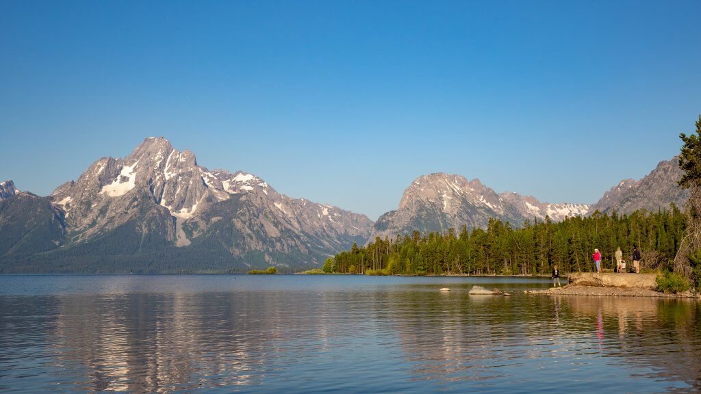 A lake lined by trees on the right side and the Teton Range in the distance.