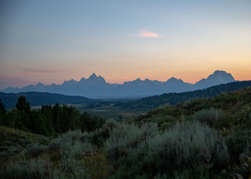 A wide shot of a landcape covered with brush and trees and the Teton Range in the distance at Grand Teton National Park.