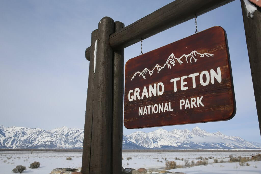 A wooden sign that reads Grand Teton National Park in the foreground and a large, open field of snow and the snow-covered Teton Range in the background.