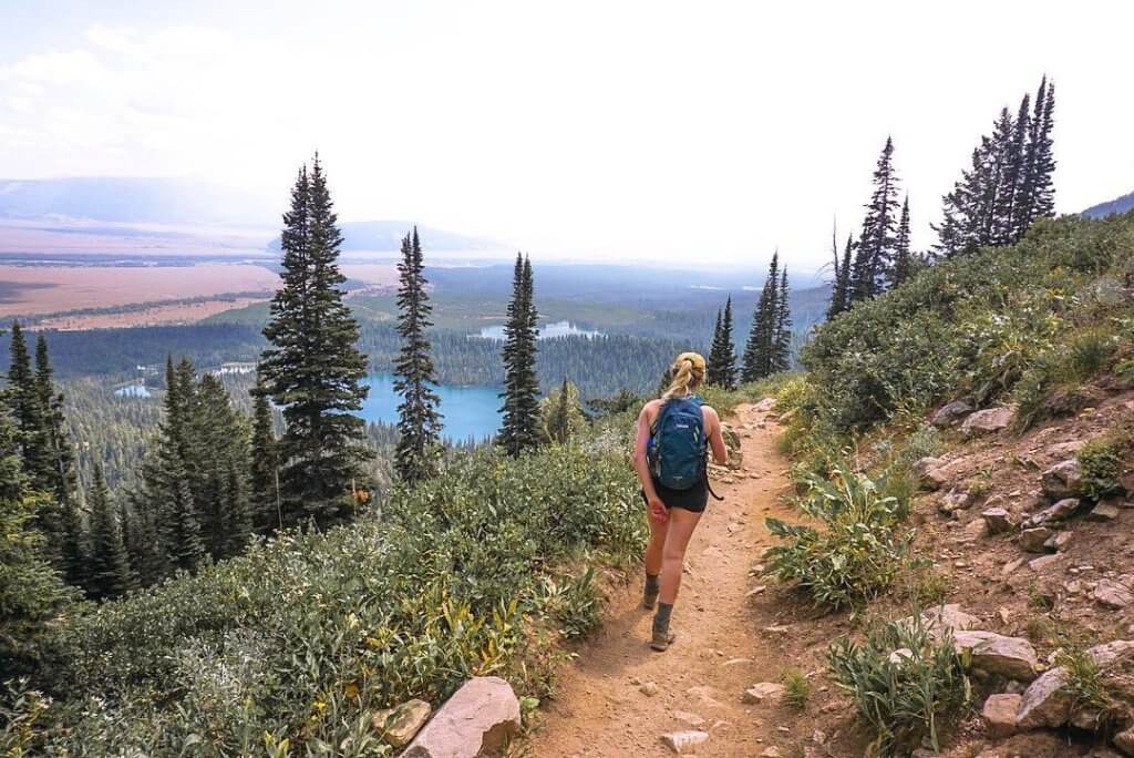 A woman hiking a long a mountain trail with a forest of trees and bodies of water in the distance at Grand Teton National Park.