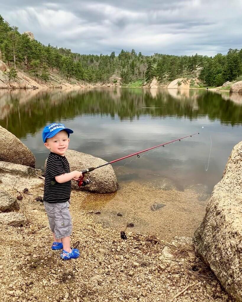 A happy young boy wearing a baseball cap holds a fishing rod, ready to cast into the waters of Curt Gowdy State Park in Laramie County, Wyoming, as trees reflect in the water.