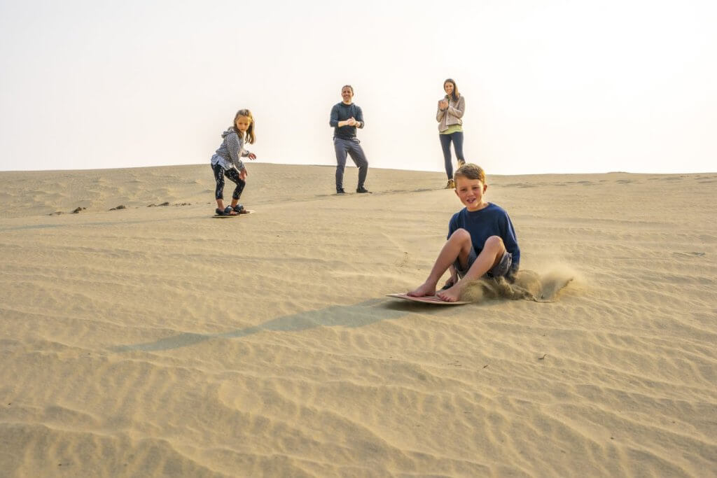 A young boy sits on a sandboard as grains of sand jump, his sister balancing on another sandboard behind him as their parents smile and clap, watching their children learn to sandsurf on the dunes. 