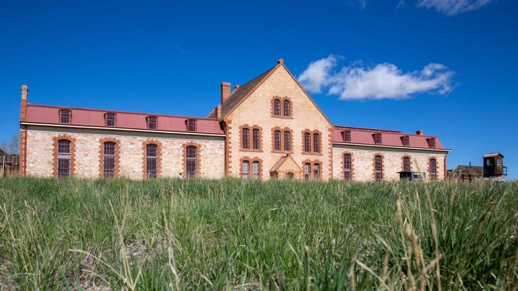 The grass sways before the Wyoming Territorial Prison State Historic Site as a wisp of cloud moves above in a bright blue sky. 