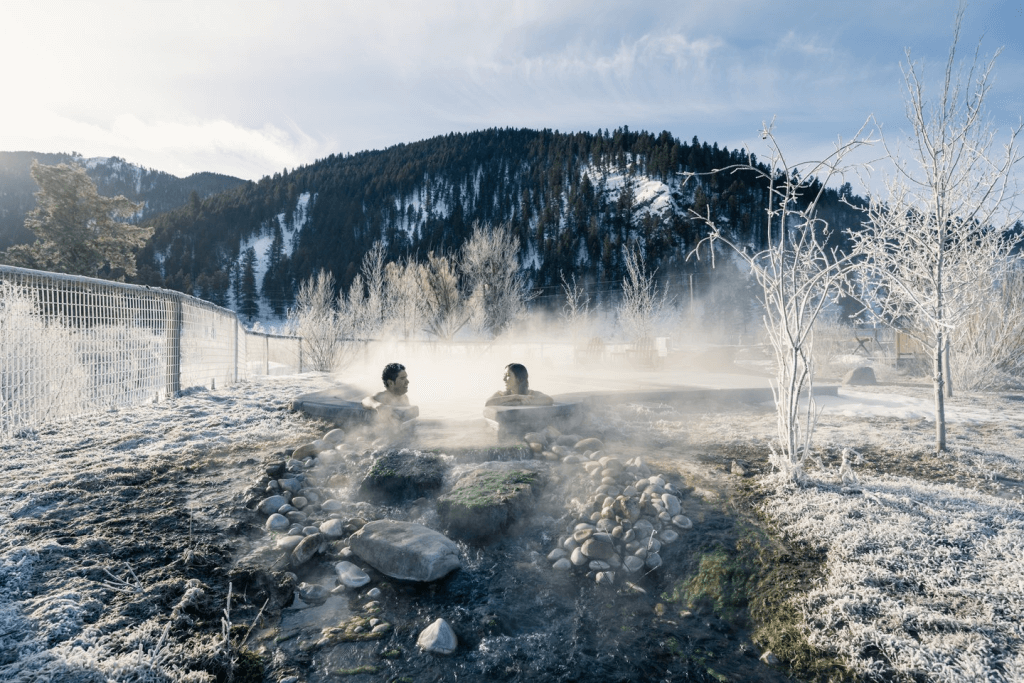 A couple takes a misty winter soak in the natural hot springs in Wyoming, surrounded by stones and frozen trees as steam rises from the icy ground.